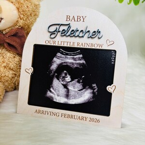 May include: A light wooden frame displaying a sonogram image, with the text "BABY Fletcher OUR LITTLE RAINBOW" and "ARRIVING FEBRUARY 2026". The frame features heart-shaped embellishments and a curved top, with a soft, natural wood finish.