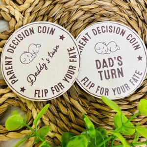 May include: Two circular wooden tokens with brown text. One reads "PARENT DECISION COIN" and "Daddy's turn", the other reads "PARENT DECISION COIN" and "DAD'S TURN". Both say "FLIP FOR YOUR FATE". A baby graphic is in the center.
