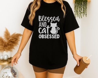 Może przedstawiać: Czarny t-shirt z białym napisem "Blessed and cat obsessed", z sylwetką kota. Koszulka ma swobodny styl, odpowiedni do noszenia na co dzień. Modelka ma na sobie brązowe szorty.