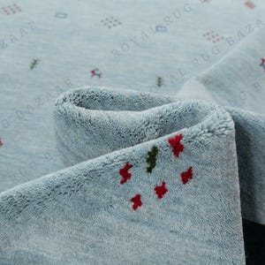 May include: A close-up of a light blue rug with a rolled edge. The rug has a soft texture and is decorated with small, scattered red and green floral patterns. The rug is a home decor item.
