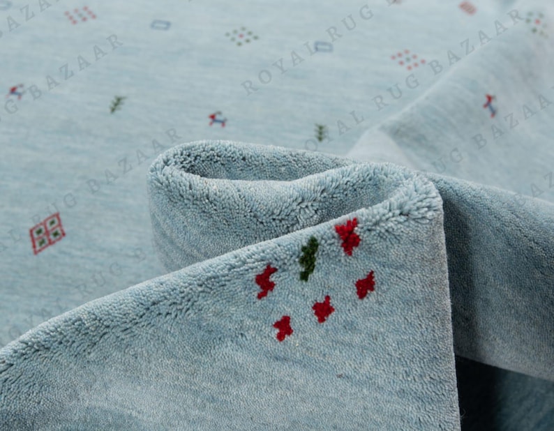 May include: A close-up of a light blue rug with a rolled edge. The rug has a soft texture and is decorated with small, scattered red and green floral patterns. The rug is a home decor item.