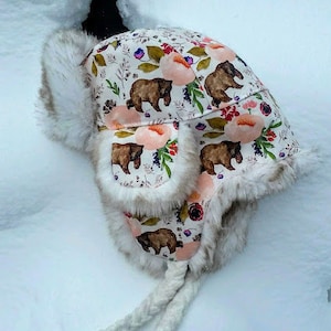 Puede incluir: Un gorro de invierno con un estampado floral y de oso. El gorro presenta un fondo blanco con osos marrones, flores rosas y hojas verdes. Tiene ribete de piel sintética y orejeras, con lazos trenzados.