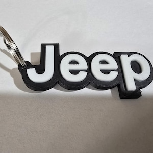 May include: A black and white Jeep logo keychain. The word "Jeep" is in white, with a black outline, and attached to a silver key ring. The keychain is on a white surface.