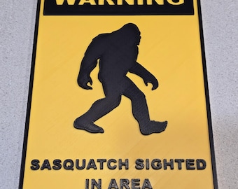 May include: A rectangular sign with a black border, yellow background, and black text. The top text reads "WARNING" above a black silhouette of a Sasquatch. The bottom text reads "SASQUATCH SIGHTED IN AREA".
