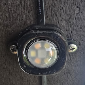 Puede incluir: Primer plano de una luminaria LED negra con una lente clara y redondeada. La luz está montada sobre una superficie de madera oscura con dos tornillos. Un cable negro está conectado a la luminaria.