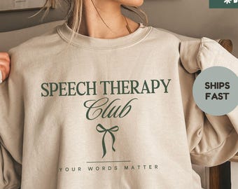 May include: Sand-colored sweatshirt with the text "SPEECH THERAPY Club" in dark green, along with a bow graphic. The phrase "YOUR WORDS MATTER" is below the club name. The sweatshirt also has the text "SHIPS FAST" and "SAND".