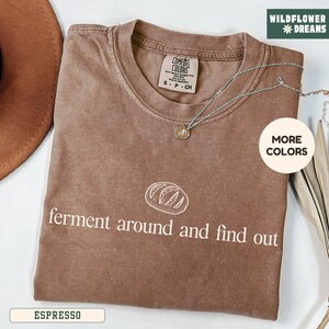May include: Espresso-colored t-shirt with the text "ferment around and find out" and a bread graphic. The shirt has a crew neck and short sleeves. A silver necklace with a pendant is draped across the shirt.