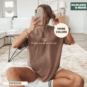 May include: Espresso-colored t-shirt with the text "ferment around and find out" in a white, stylized font. The shirt is a solid color and has a crew neck. The image also includes the words "MORE COLORS" and the brand name "WILDFLOWER DREAMS".