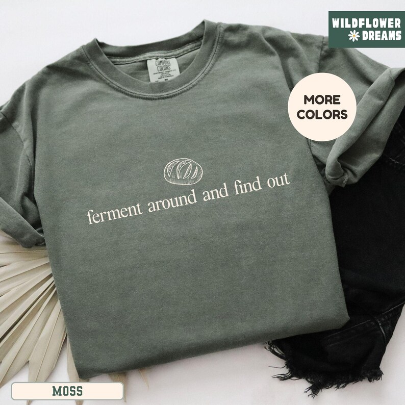 May include: A moss green t-shirt with the text "ferment around and find out" in white. The shirt features a simple bread graphic above the text. The shirt is displayed with black denim jeans and a white background.