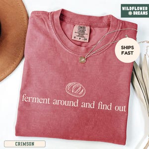 May include: Crimson-colored t-shirt with the text "ferment around and find out" and a bread graphic. The shirt has a crew neck and a small tag. A silver necklace with a pendant is draped across the shirt.