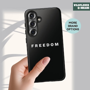 May include: Black phone case with the word "FREEDOM" in white block letters. The phone case has a triple-lens camera and the "Wildflower Dreams" logo in the upper right corner. The words "MORE BRAND OPTIONS" are in a circle.