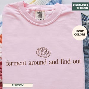 May include: A light pink t-shirt with the text "ferment around and find out" and a bread graphic. The shirt has a crew neck and a tag that says "Comfort Colors". The shirt is folded on a surface.