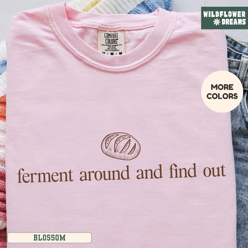 May include: A light pink t-shirt with the text "ferment around and find out" and a bread graphic. The shirt has a crew neck and a tag that says "Comfort Colors". The shirt is folded on a surface.