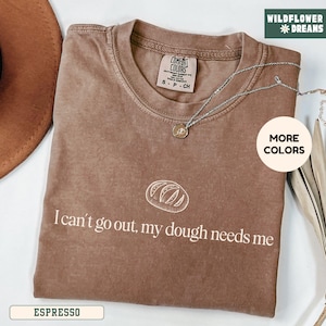 May include: Espresso-colored t-shirt with the text "I can't go out, my dough needs me" and a bread graphic. The shirt has a crew neck and short sleeves. A silver necklace with a pendant is draped across the shirt. The shirt is labeled "Comfort Colors."