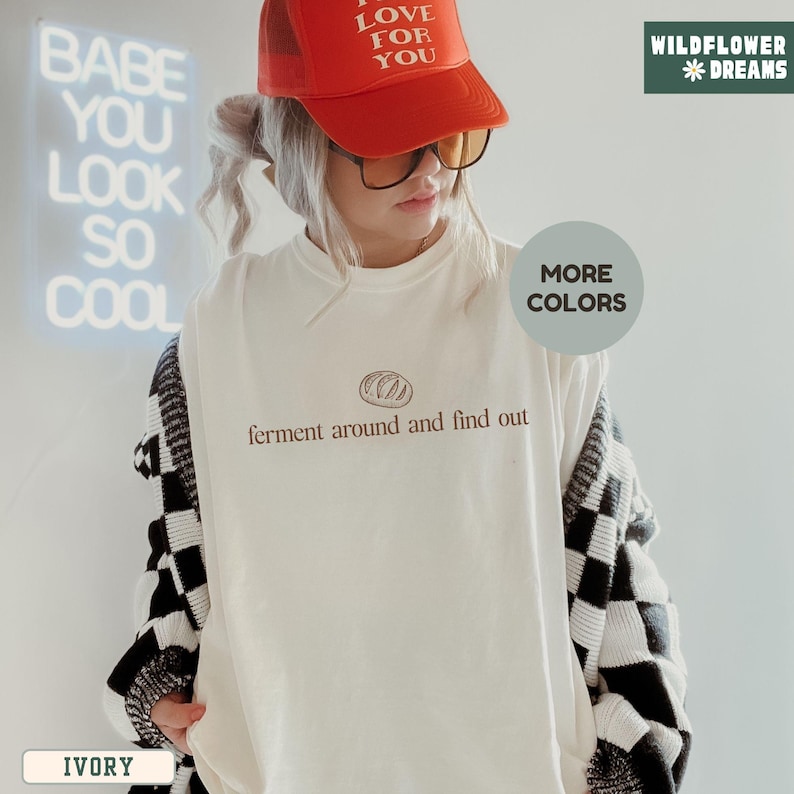 May include: An ivory-colored t-shirt with the text "ferment around and find out" in brown. The person is wearing a red trucker hat with "I love for you" printed on it, and a black and white checkered cardigan. The background has a neon sign that says "Babe you look so cool".