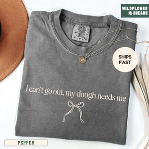 May include: A gray Comfort Colors t-shirt with the text "I can't go out, my dough needs me" and a bow graphic. A silver necklace with a pendant is draped across the shirt. The shirt is labeled "Pepper".