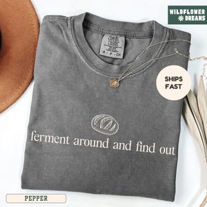 May include: Pepper-colored t-shirt with the text "ferment around and find out" and a bread graphic. The shirt has a crew neck and short sleeves. A silver necklace with a pendant is draped across the shirt.