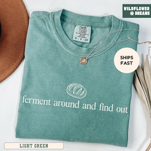 May include: Light green t-shirt with the text "ferment around and find out" and a bread graphic. The shirt has a crew neck and short sleeves. A silver necklace with a pendant is draped across the shirt. The shirt is labeled "Comfort Colors".