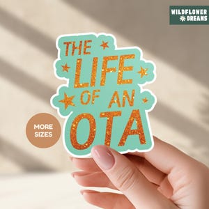 May include: A sticker with the text "THE LIFE OF AN OTA" in orange glitter on a teal background. The sticker has a white border and star accents. A small brown circle with the words "MORE SIZES" is on the left.