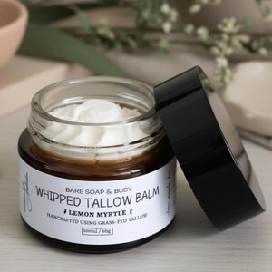 May include: A glass jar of whipped tallow balm with a black lid. The jar has a white label with the text "WHIPPED TALLOW BALM" and "LEMON MYRTLE". The jar contains a white, creamy substance. The product is handcrafted using grass-fed tallow. The jar contains 100ml / 60g.