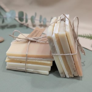 May include: Several stacks of handmade soap bars in shades of cream, beige, and light brown, bound with twine. Each bar is rectangular and has a smooth surface. A small tag with the word "Handmade" is attached.