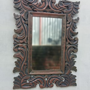 May include: Ornate rectangular mirror with a dark brown, carved wooden frame. The frame features a detailed, swirling leaf design. The mirror is mounted on a white wall, reflecting a blurred outdoor scene.