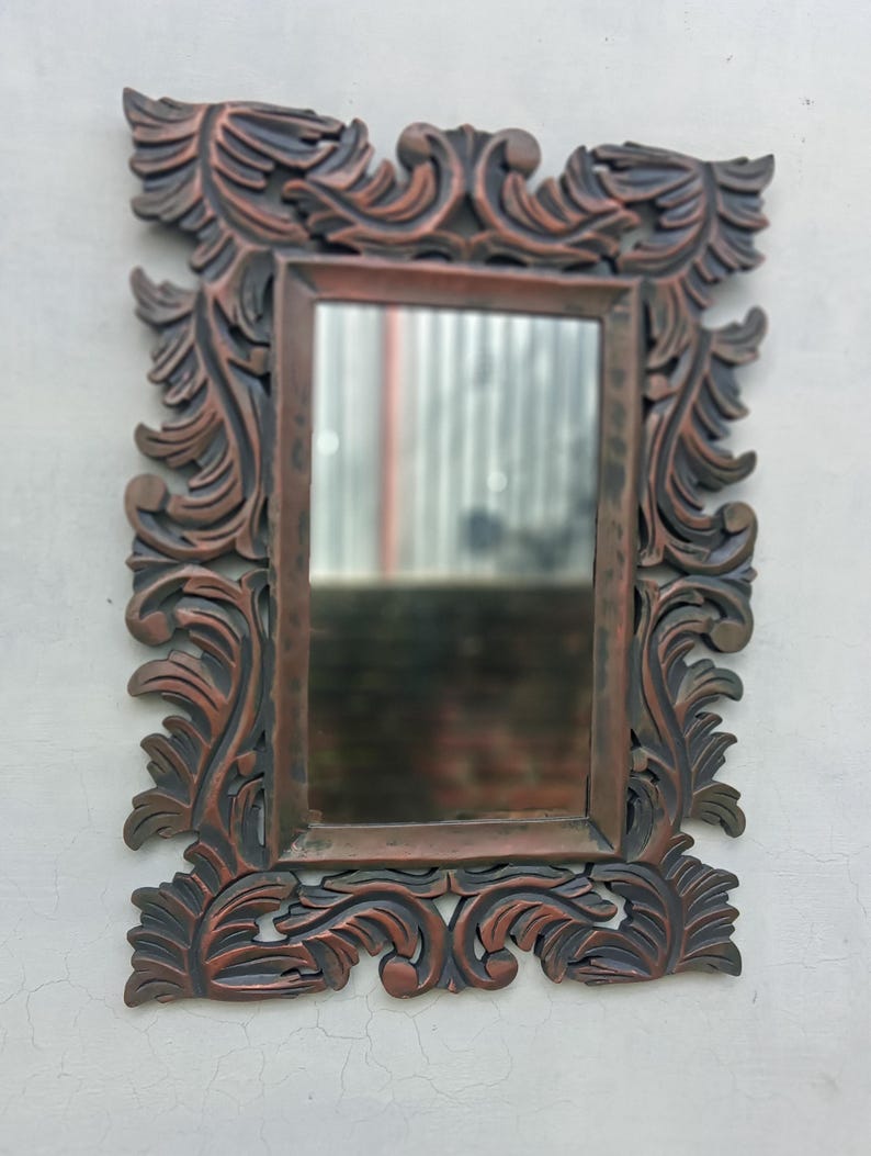 May include: Ornate rectangular mirror with a dark brown, carved wooden frame. The frame features a detailed, swirling leaf design. The mirror is mounted on a white wall, reflecting a blurred outdoor scene.