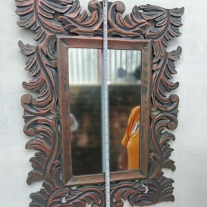 May include: Ornate rectangular mirror with a dark brown and copper-colored carved frame. The frame features a detailed, swirling leaf design. The mirror's height is approximately 30 inches.