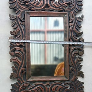 May include: Ornate rectangular mirror with a dark brown, carved wooden frame. The frame features detailed, swirling leaf and scroll designs. The mirror's dimensions are approximately 20 inches wide.