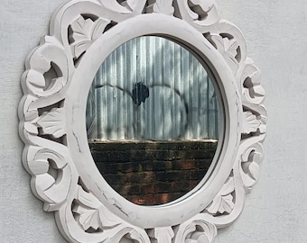 May include: Oval white mirror with ornate, carved floral detailing. The mirror has a distressed finish, and the frame surrounds a circular reflective surface. The mirror is mounted on a textured, light-colored wall.