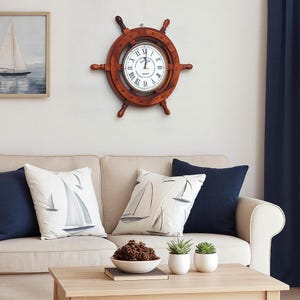 Puede incluir: Un reloj de rueda de barco de madera con esfera blanca y números romanos cuelga sobre un sofá beige con cojines azul marino y cojines con temática de veleros. Una pintura enmarcada de un velero está en la pared.