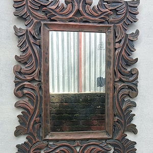 May include: Ornate rectangular mirror with a dark brown, carved wooden frame. The frame features detailed, swirling leaf and scroll designs. The mirror reflects a view of a corrugated metal surface and a brick wall. The mirror is mounted on a white wall.
