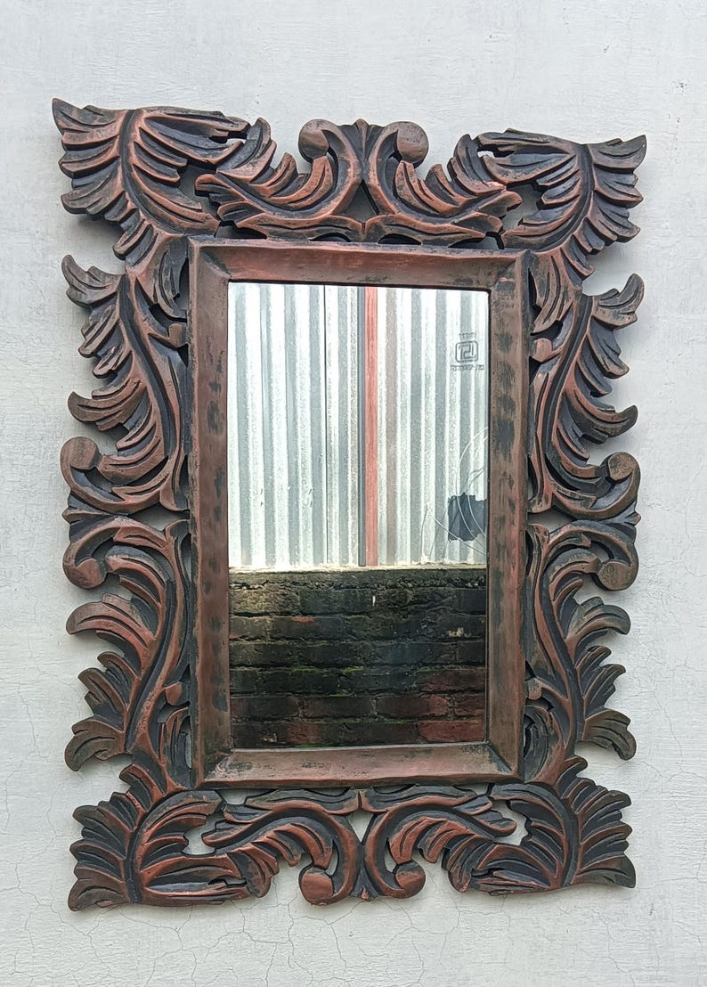 May include: Ornate rectangular mirror with a dark brown, carved wooden frame. The frame features detailed, swirling leaf and scroll designs. The mirror reflects a view of a corrugated metal surface and a brick wall. The mirror is mounted on a white wall.