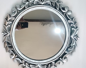 May include: A round silver mirror with an ornate floral frame. The frame features detailed leaf and scroll designs. The mirror's reflective surface shows a partial reflection. The mirror is designed for wall mounting and adds a decorative touch to any room.