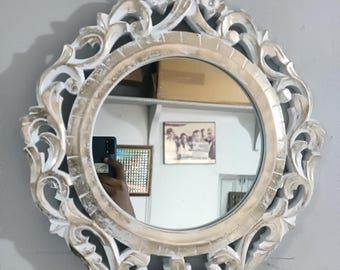 Op de afbeelding: Een ronde, sierlijke spiegel met een crème-kleurig, gesneden houten frame. Het frame heeft bloemen- en krulwerkontwerpen. De spiegel reflecteert een kamer met diverse objecten. De spiegel is op een grijze muur gemonteerd.