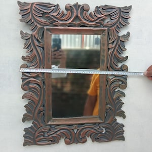 May include: Ornate rectangular mirror with a bronze-colored, carved frame. The frame features detailed floral and leaf designs. The mirror measures approximately 20 inches wide. The mirror reflects a person holding a measuring tape.