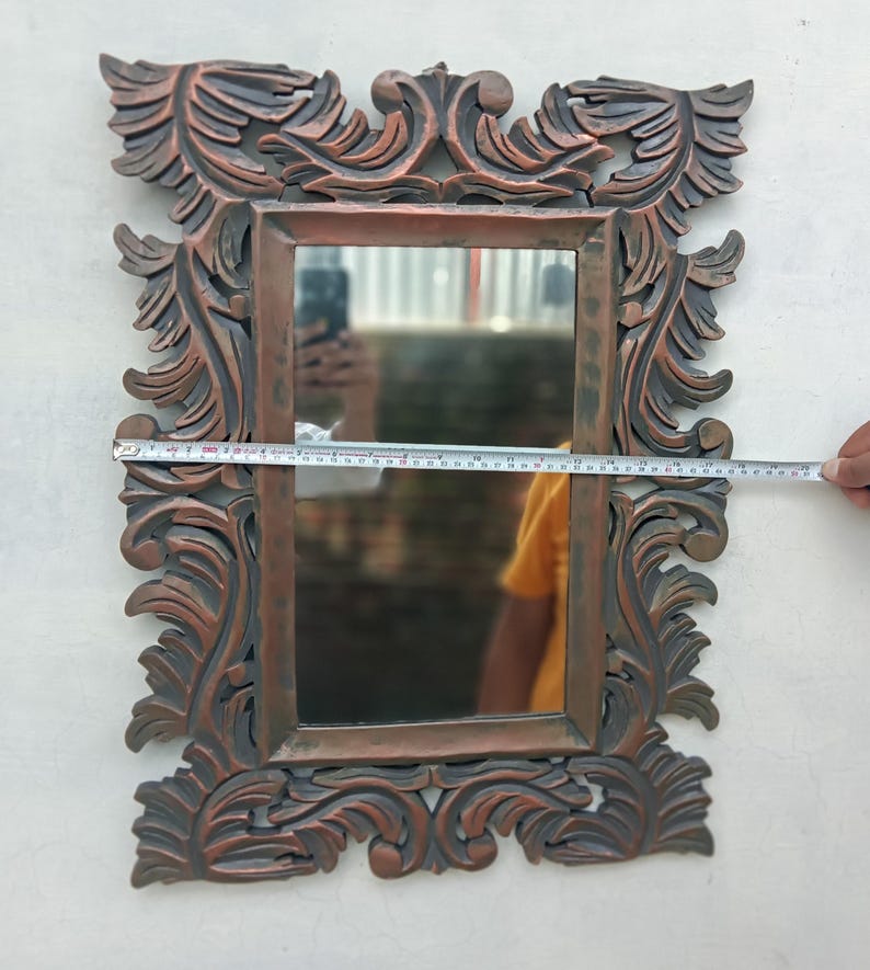 May include: Ornate rectangular mirror with a bronze-colored, carved frame. The frame features detailed floral and leaf designs. The mirror measures approximately 20 inches wide. The mirror reflects a person holding a measuring tape.