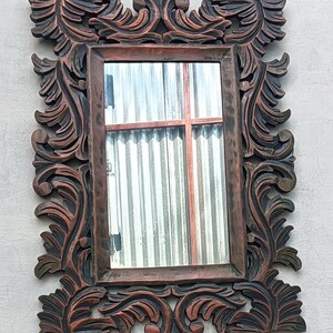 May include: Ornate rectangular mirror with a dark brown, carved wooden frame. The frame features detailed, swirling leaf and scroll designs. The mirror reflects a window with corrugated metal siding and a red cross-shaped structure.