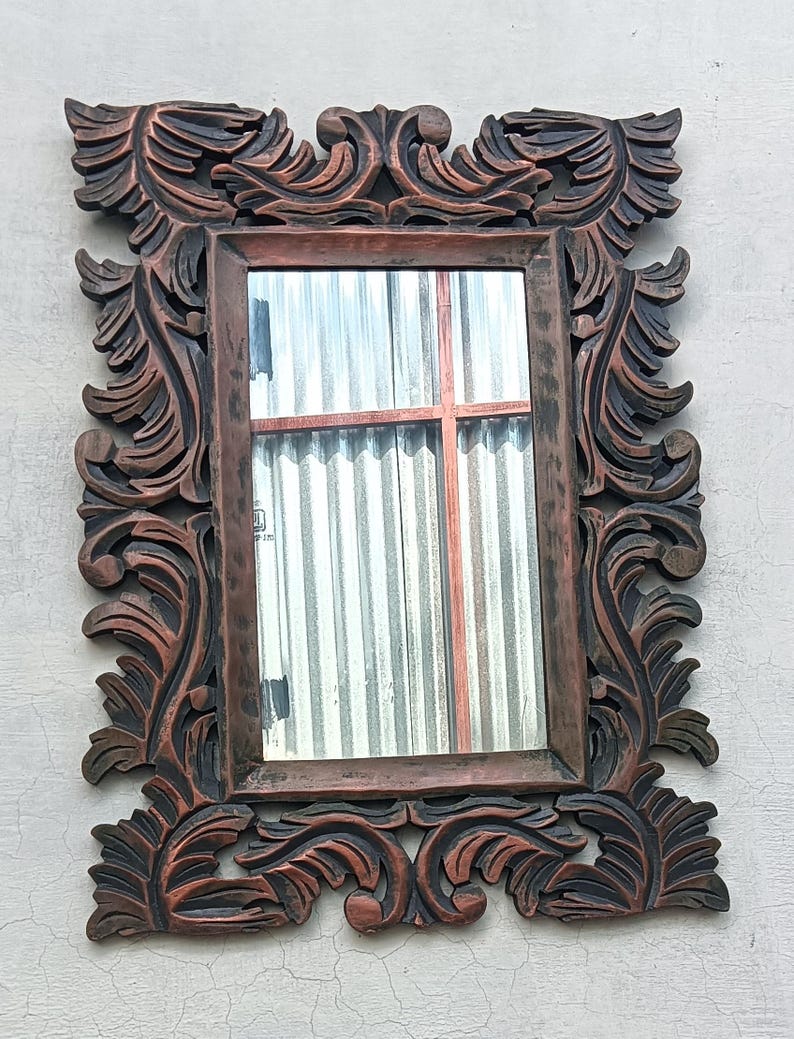 May include: Ornate rectangular mirror with a dark brown, carved wooden frame. The frame features detailed, swirling leaf and scroll designs. The mirror reflects a window with corrugated metal siding and a red cross-shaped structure.