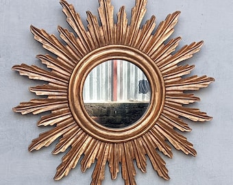 May include: A circular wall mirror featuring a gold-toned frame designed with radiating, leaf-like rays extending outward from the center. The reflective glass surface is surrounded by a smooth, rounded inner border and a textured, sunburst-style edge.