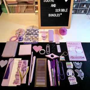 May include: An assortment of purple stationery items, such as pens, sticky notes, a journal, and decorative paperclips, arranged on a black surface. A letter board displays the words "SOOTHE AND SCRBBLE BUNDLES".