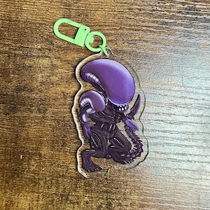 以下が含まれることがあります： 緑色の留め金が付いた、紫と黒のエイリアンのキーホルダー。キーホルダーには、映画「エイリアン」シリーズに登場するゼノモーフの生き物の詳細なイラストが描かれています。キーホルダーは透明なアクリル製です。