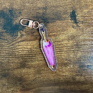 Peut inclure: Un porte-clés en acrylique transparent avec un design de flacon de potion violet et des accents dorés. Le porte-clés a un fermoir doré et une petite gemme violet foncé en haut. Le flacon de potion a une base pointue.