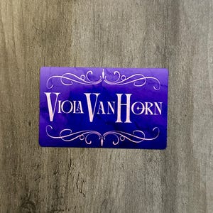 Op de afbeelding: Een rechthoekige kaart met de tekst "Viola Van Horn" in witte, gestileerde letters. De kaart heeft een paarse tot blauwe gradiënt achtergrond. Witte decoratieve krullen en krullen sieren de boven- en onderkant.