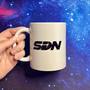 May include: A white ceramic mug with a black logo that reads "SDN" with a star design. The mug has a curved handle and is held by a hand. The background is a blue and purple galaxy design.