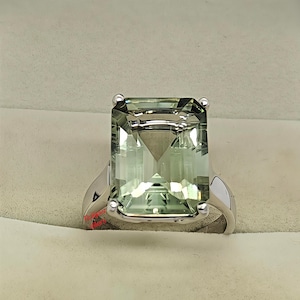 May include: A silver ring featuring a large, rectangular-cut, pale green gemstone. The gemstone is held in place by four prongs. The ring is presented on a light-coloured surface.