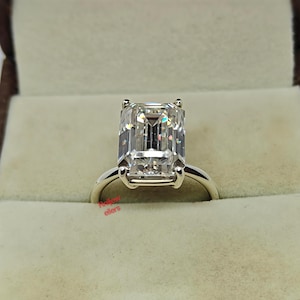 Peut inclure: Une bague de fiançailles en argent avec un grand diamant de taille émeraude rectangulaire. La bague est sertie dans un sertissage classique à quatre griffes, et le bandeau est lisse et poli. La bague est présentée dans un écrin de couleur claire.