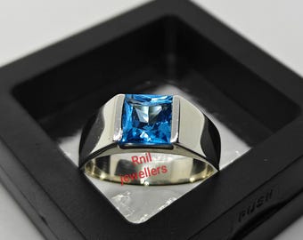 May include: A silver ring with a large, square, bright blue gemstone. The ring is set in a simple, modern style, with a wide band. The ring is displayed in a black box, highlighting the jewelry's color and design.