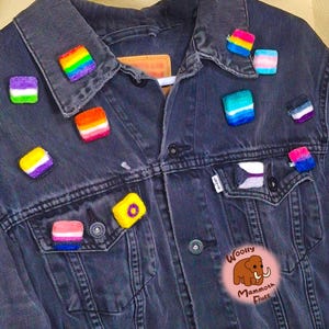 May include: A dark gray denim jacket adorned with multiple small, square, fuzzy pins featuring various colorful striped patterns. A circular pink logo with a brown mammoth illustration and the text Woolly Mammoth Fluff is visible.
