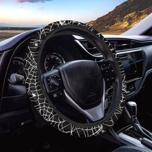 May include: Black steering wheel cover featuring a white spiderweb and spider design. The cover is fitted on a black steering wheel inside a car. The background shows a dashboard and a mountain range.